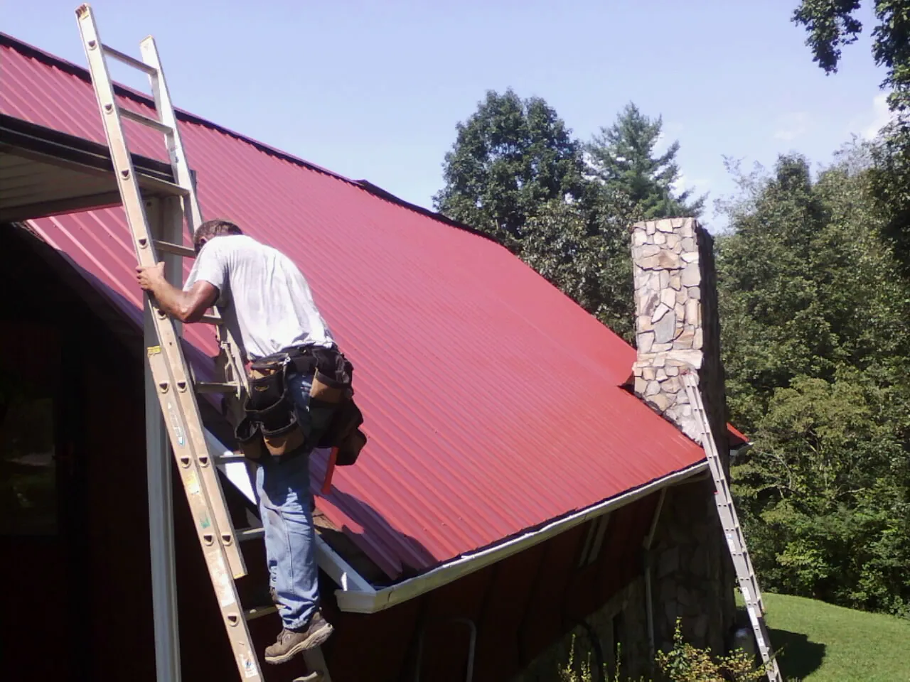 Roofer on a ladder beside a red metal roof and stone chimney — Soffit & Fascia Repair by Roofing Brothers in Cornersville